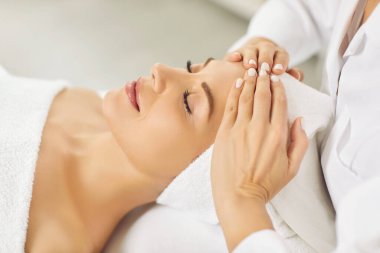 Kapalı gözlerle uzanan ve kaplıca salonunda yüz veya kafa masajı yaptıran güzel bir genç kadının yan görüntüsü. Kadın, rahatla ve sağlık terapisinin tadını çıkar. Sağlık ve güzellik günü konsepti.