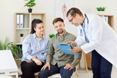 Genç ve mutlu bir çift, klinik planlama muayenesi sırasında randevu almak isteyen bir erkek aile doktoru ile konuşuyor. Sağlık hizmeti kavramı.