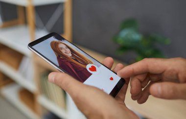 Çevrimiçi aşk. Kapanış adamı kırmızı kalp tuşuna basıyor ve güzel kızıl saçlı genç kızların flört sitesindeki veya cep telefonu uygulamasındaki resimlerine benziyor. Eller cep telefonunu yakın çekimde tutsun.