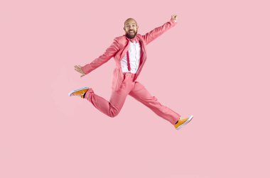 Mutlu, takım elbiseli genç adam pastel pembe arka planda izole bir şekilde atlarken eğleniyor. Pembe takım elbiseli, pantolon askısı, papyon ve spor ayakkabısı olan beyaz bir adam kameraya bakıyor. Pankart. Tam uzunluk.