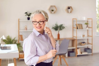 Arkadaş canlısı kıdemli iş kadını ofisinde cep telefonuyla iş görüşmesi yapıyor. Gülümserken kulağına akıllı telefon dayayan zarif, olgun bir kadının portresi..