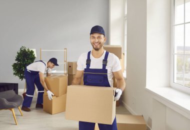 Portrede gülümseyen bir nakliyeci bir ofis ya da ev taşıma sırasında kutu tutuyor. Davranışları, bir profesyonelin, dostça bir dokunuşla nakliye hizmetleri sunmasını yansıtıyor..