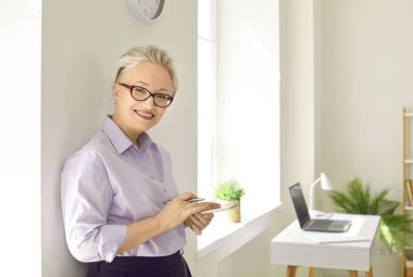 Başarılı, gülümseyen, cep telefonlu yaşlı kadın, kameraya kendine güvenen, şık, resmi giysiler giymiş, kendi ofisindeki pencerenin yanında duran,