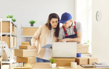 Depoda birlikte çalışan bir adam ve kadın, paket ve paketlerin envanterini çıkarmak için dizüstü bilgisayar kullanıyorlar. Takım çalışması, verimlilik ve iş operasyonları ve depolama alanında teknolojinin kullanımı.