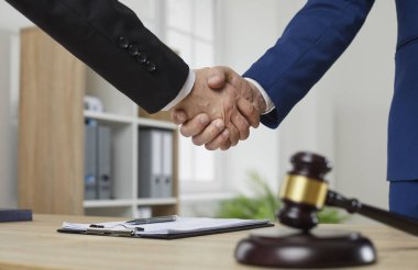 Profesyonel avukat müşteriyle tokalaşıyor. Takım elbiseli iki adam ofis masasının üzerinde yargıç tokmağı ile tokalaşıyor. İnsan elinin yakın plan çekimi. Hukuk danışmanlığı, hukuk hizmetleri kavramı