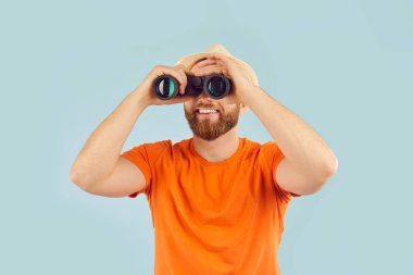 Mutlu gülümseyen genç turist portresi günlük kıyafetler ve plaj şapkası giymiş stüdyo mavi arka planında dürbünle bakan. Yaz tatili gezisi, arama ve tatil konsepti.