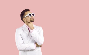 Düşünceli ifadeli şık, havalı bir genç adam plan yapmayı düşünüyor ya da aklında bir fikir var. Beyaz ceketli, güneş gözlüklü, çenesine dokunup fotokopi çeken beyaz bir adam. Pembe arka planda izole edilmiş.