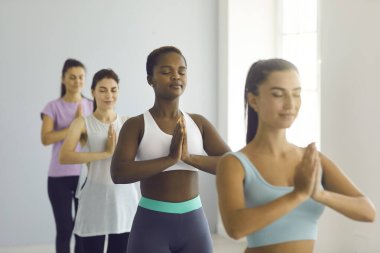 Grup Meditasyonu. Rahat Afro-Amerikalı genç bir kadın grup yoga sınıfında kollarını kavuşturup namaste jesti yapıyor. Kadın, beyaz kadınlarla birlikte parlak bir salonda meditasyon yapıyor. Seçici odak.