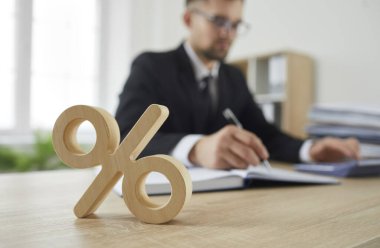 Şirket vergisi ve faiz oranı sembolü olarak masadaki ahşap yüzde işaretini kapat. Takım elbiseli, hesap makinesinde hesap yapan ciddi meşgul bir adamın arka planında duran yüzde işareti. Bulanık arkaplan.