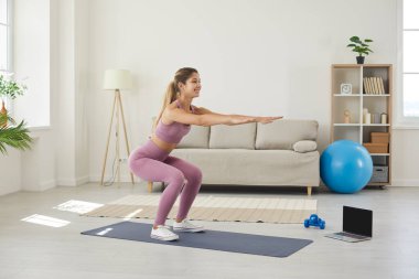 Sanal fitness hocasıyla evde antrenman yapan mutlu bir kadın. Oturma odasında yoga minderinin üzerinde duran ve boş ekranla laptopun yanında çömelme egzersizi yapan genç, beyaz bir bayan.