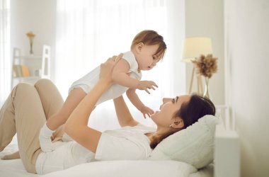 Sevinçten havalara uçmuş genç bir anne evde yatıp yeni doğmuş küçük bir bebekle oynuyor. Sevgi dolu, mutlu, beyaz bir anne, küçük bir çocukla birlikte neşeli bir rahatlama hisseder. Annelik zamanı konsepti.