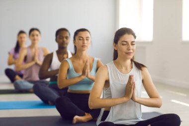 Yoga stüdyosunda meditasyon dersi alan bir grup güzel genç kadın. Çok ırklı kadınlar spor salonlarında oturuyor, namaste jestleri yapıyor, gözleri kapalı meditasyon yapıyor ve enerji seviyelerini yükseltiyorlar.