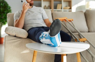 Bacağında bandaj olan yaralı bir adama yaklaş. Kanepeye otur ve doktorla cep telefonundan konuş. Kırık ayaklı sağlıksız bir adam kırık ya da yaralanmadan muzdarip. Ev konseptinde rehabilitasyon.