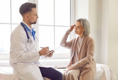 Doktor danışmanlık kadın kıdemli hasta belirtileri paylaşıyor, memnuniyetsizlik belirtileri gösteriyor, kafa ve hafıza rahatsızlığı gösteriyor. Yaşlı kadın tıbbi bakım için kliniğe gidiyor, tedavi planını tartışıyor, yardım istiyor.
