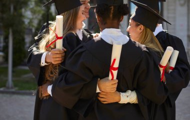 Mutlu gülümseyen çeşitli öğrenci grupları açık havada diplomaları ellerinde tutarak mezuniyetlerini kutluyorlar. Üniversite kampüsünde mezuniyet elbisesi içinde halka halinde duruyorlar..