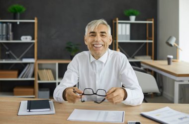 Beyaz gömlekli yetişkin bir adam büro masasında oturuyor, elinde gözlük tutuyor ve mutlu bir yüz ifadesiyle kameraya bakıyor. Gülümseyen kıdemli işadamı, işletme yöneticisi, CEO ya da mali danışman portresi