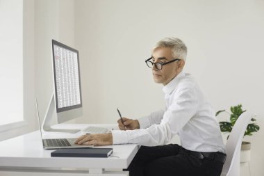 Modern ofiste çalışan olgun muhasebeci iş adamı. Gözlüklü ciddi bir beyaz adam dizüstü bilgisayar ve bilgisayarla masada oturuyor, elektronik iş kağıtlarıyla çalışıyor ve notlar alıyor.