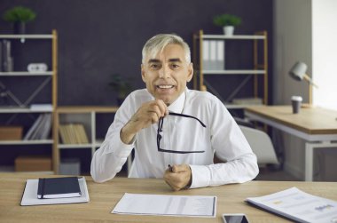Ofis masasında oturan, elinde gözlük olan ve kameraya bakan son sınıf öğrencisinin portresi. Olgun iş adamı, CEO, yönetici, finans danışmanı, iş adamı ya da İnsan Kaynakları müdürünün webcam görüntüsü