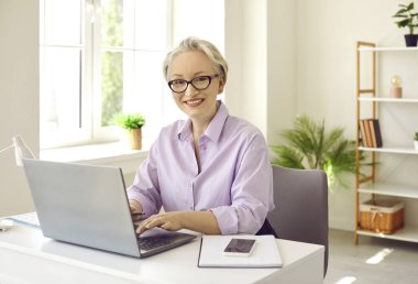 Masada oturan orta yaşlı bir kadın dizüstü bilgisayarla çalışıyor. Çekici, gri saçlı, kendine güveni olan, rahat bir ofiste çalışırken bilgisayarda yazan iş kadını.