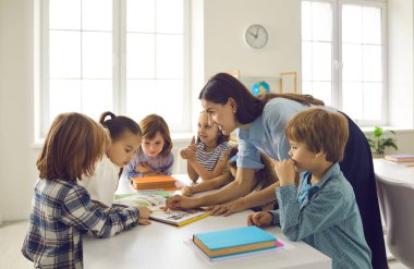 Mutlu öğretmen ve çocuklar birlikte kitap okuyor. Bir grup öğrenci, öğretmen tarafından anlatılan ilginç hikayeyi dinlemek için sınıftaki masanın etrafında toplanıyor. İlkokul öğrencileri sınıfta yeni şeyler öğreniyor.