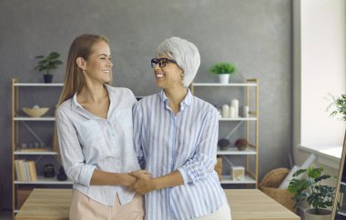 Yaşlı anneyi ve yetişkin kızı birlikte sevmenin aile portresi. Farklı yaşlarda iki kadın masanın yanında durup birbirlerine bakıyorlar ve sağlıklı, güzel, pozitif beyaz gülücüklerle gülümsüyorlar.