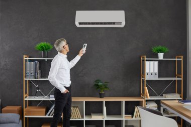 Orta yaşlı beyaz adam modern ev oturma odasındaki klimayı çalıştırıyor. İş adamı ya da CEO temiz hava almak için modern elektronik havalandırma cihazını değiştirdi. Teknoloji konsepti.