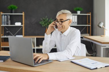 İşyerinde otururken bilgisayarla çalışan ve cep telefonuyla konuşan neşeli kıdemli işadamı. Arkadaş canlısı yönetici, müşteriye telefonla danışıyor. Uzak çalışma kavramı.