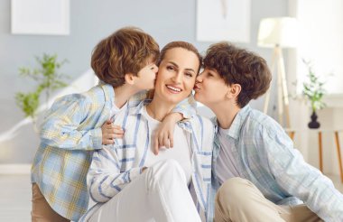 Mutlu bir anneyi öpen iki çocuğun portresi. Oturma odasında yerde oturuyor. Genç kadın ebeveyn, oğullarıyla vakit geçirmekten hoşlanıyor. Aile eğlencesi ve anneler günü konsepti.