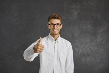 Gözlüklü mutlu adam, koyu gri arka planda tek başına ayakta duruyor. Yakışıklı yetişkin öğrenci portresi harika bir fikri onaylıyor, jest yapıyor ya da el işareti yapıyor.