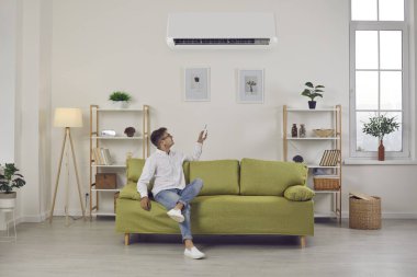 Klimaları değiştiren bir adam evde temiz havanın tadını çıkarırken rahat bir sıcaklık yaratıyor. Uzaktan kumandalı bir adam oturma odasında kanepede oturuyor. Klima sistemi kavramı.