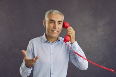 Kıdemli adam sabit kırmızı hattan biriyle konuşuyor. Ciddi yakışıklı, yaşlı bir adamın portresi gri stüdyo arka planında duruyor, elinde retro telefon alıcısı, konuşuyor ve el kol hareketi yapıyor.