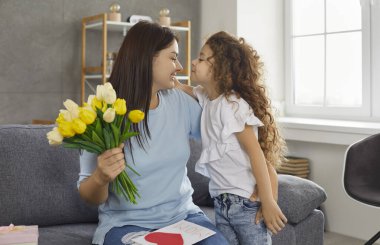 Mutlu beyaz kadın, Anneler Günü 'nde sevgi, şefkat ve çocuğundan hediyeler alıyor. Annem evdeki kanepede oturuyor. Eskimo tatlı küçük kızıyla öpüşüyor ve çiçekler ve kartpostallar için ona teşekkür ediyor.