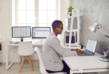 Ofiste dizüstü bilgisayar kullanan siyah bir kadın. Mutlu genç muhasebeci ya da iş kadını, modern yazılımlar kullanarak, dijital arşivler ve hesap tablolarıyla çalışıyor.