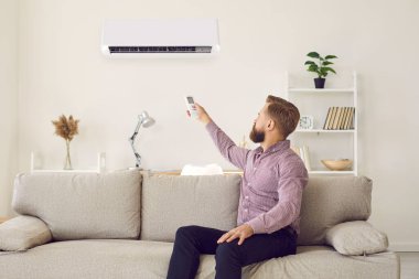 Erkek kiracı rahat bir kanepeye uzanıp evdeki temiz hava için uzaktan kumandalı klimayı çalıştırır. Kiracı erkek kanepede dinleniyor. İçerideki havalandırma elektronik cihazını kullan..