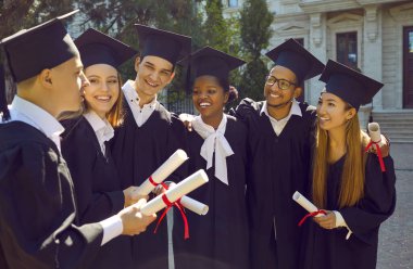 Neşe dolu, başarılı, çok ırklı mezunların açık havada ellerinde diplomalarla portresi. Havan ve manto taşıyan gülümseyen öğrenciler sıraya girip diplomalarını yükseltiyorlar..