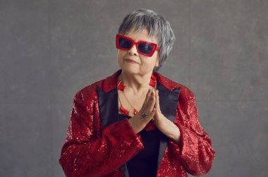 Gri stüdyo arka planında duran kırmızı pullu disko ceketli ve havalı güneş gözlüklü yaşlı gri saçlı kadın. Şık giyinmiş yaşlı ninenin portresi parti için hazır.