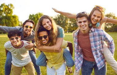 Yaz yürüyüşünde eğlenen genç erkek ve kadın öğrenciler birbirlerini kollarında taşıyorlar ve tatilde dinlenen kameraya bakıp güneşli parkta poz veriyorlar.