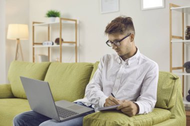 Ciddi bir genç adam çalışmak için dizüstü bilgisayar kullanıyor. Gözlüklü üniversite öğrencisi evdeki kanepede oturuyor, online ders alıyor, bilgisayar ekranına bakıyor ve not defterine not alıyor. Uzaklık öğrenme kavramı