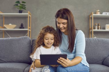 Evde modern tablet kullanan mutlu bir aile. Genç anne ve sevimli küçük kız gülümsüyor, cihaz ekranına bakıyor, akrabalarını arıyor, konuşuyor ya da eğlenmek için iyi bir film ya da çizgi film programı seçiyor.