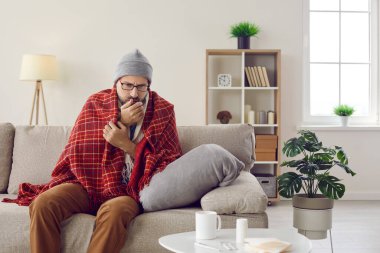 Sağlıksız bir adam evinde battaniyenin içinde oturur ve kovid 19 Corona virüsüyle buz gibi bir mücadele yaşar. Hasta erkek öksürüğünün ateşle ya da griple boğuştuğu için yüksek ateşi var. Coronavirus konsepti.