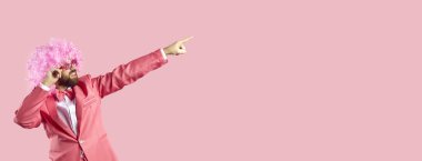 Neşeli adamın portresi sağdaki boş metin arkaplanına işaret ediyor. Komik pembe takım elbiseli komik palyaço, kıvırcık karnaval peruğu ve güneş gözlüğü sizi havalı bir partiye ya da harika bir tatil indirimine davet ediyor.