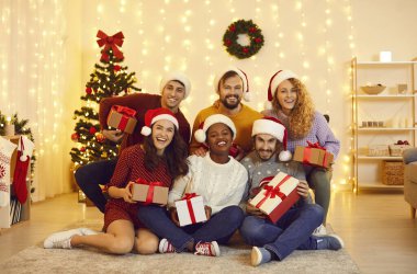 Yeni yılı evde birlikte kutlayan mutlu genç arkadaşların toplu portresi. Noel Baba şapkalı çok ırklı insanlar ellerinde hediye kutuları ve gülümseyerek odada Noel dekoruyla kameraya bakarlar..
