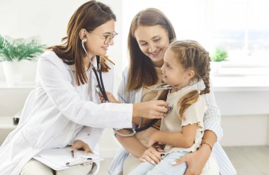 Pediyatrist muayene odasında genç ve neşeli annesiyle birlikte küçük kız hastayı muayene ediyor. Steteskoplu kadın aile doktoru hastanedeki çocukların akciğerlerini inceliyor..