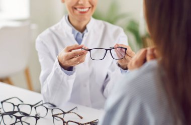 Genç güler yüzlü gözlükçü doktor masasında oturuyor ve görme düzeltmesi için klinikteki göz muayenesinden sonra hastasına yeni bir gözlük veriyor. Oftalmoloji, göz sağlığı konsepti.