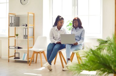 Modern ofis içinde koltuklarda oturan iki genç Afrikalı Amerikalı kadın birlikte dizüstü bilgisayar kullanıyorlar, iş projesi üzerinde çalışıyorlar ve online araştırma yapıyorlar.