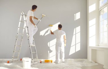 Beyaz elbiseli profesyonel ressamlardan oluşan bir ekibin boş bir odada boya silindiriyle gri bir duvar boyadığını gördüm. Müteahhitler müşteri evinde tadilat yapıyor. Onarım kavramı.