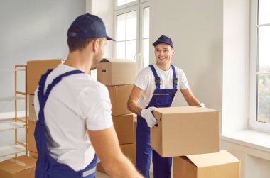 Taşıma servisi. Ulaşım ve teslimat şirketinin neşeli çalışanları karton kutuları birlikte taşır. Tulum giymiş erkek yükleyiciler boş ofiste karton kutular paketlerken neşeli bir şekilde konuşuyorlar..