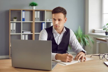 İş adamı iş programını planlıyor. Ofis masasında oturan genç adam dizüstü bilgisayara bakıyor, bazı tarihleri kontrol ediyor ve iş planlayıcısına yazı yazıyor.