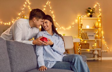 Gülümseyen adam sürpriz yapıyor, kırmızı hediye kutusu veriyor ve evdeki koltukta oturan mutlu kadını tebrik ediyor. Sevgililer Günü 'nü, yıldönümünü veya doğum gününü romantik bir ortamda kutlayan genç bir çift..