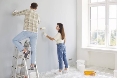 Karı ve kocanın yeni dairenin duvarlarını boyarken, ellerinde boya ruloları tutarken ve merdivenlerde dikilirken görüntüsü. Evli ve mutlu çift evlerinde tadilat ve dekorasyon yapıyor..